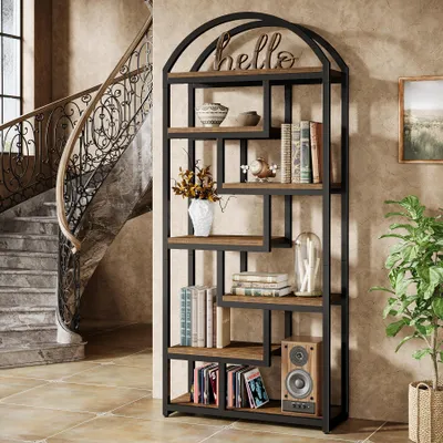 A tall, multi-shelf bookcase with a metal frame and wooden shelves, filled with books and decorative items, is placed next to a grand, curved staircase in a well-lit room.