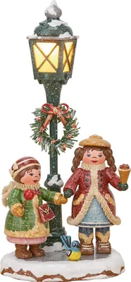 Zwei fröhliche Kinder in winterlicher Kleidung stehen neben einer Laterne mit einem Kranz und Schnee während eines von ihnen ein Eis hält und ein kleiner Vogel auf dem Boden sitzt