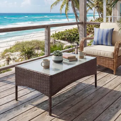 A rattan coffee table with a glass top is placed on a wooden deck overlooking a beach and the ocean.