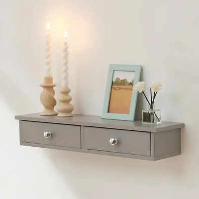 A gray floating shelf with two drawers is mounted on a white wall. It is decorated with two white twisted candles on wooden candle holders, a framed picture of a landscape, and a small glass bottle with reeds.