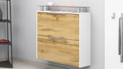A wall-mounted shoe cabinet with two drawers, featuring a white frame and wood-grain drawer fronts, placed against a light gray wall. A small white bowl and some papers are on top of the cabinet.