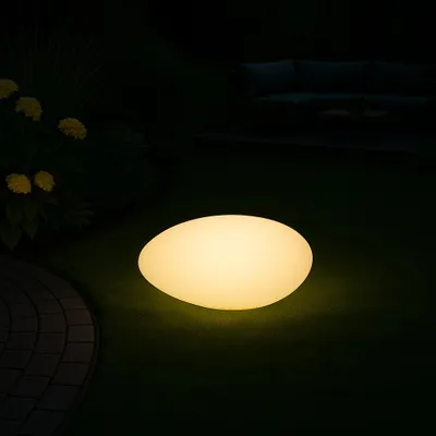 A glowing, stone-shaped garden lamp sits on a lawn at night, with some yellow flowers and a paved path visible.