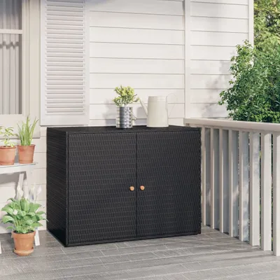 A black poly rattan garden cabinet with two doors sits on a patio next to a white railing. Plants are visible on a shelf to the left and in pots on the patio.