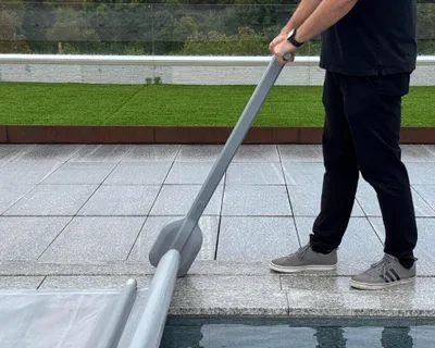 A person is using a pool cover reel to roll up a pool cover next to a swimming pool.