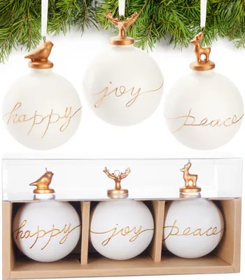 Three white Christmas ornaments with gold lettering and gold toppers are displayed hanging from a pine branch, and also in a clear plastic box with a cardboard base.
