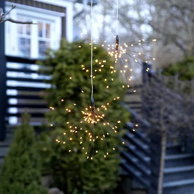 Two firework-shaped solar lights hanging outdoors in front of a house and trees.
