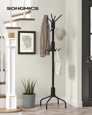 A black metal coat rack with 12 hooks stands in a hallway next to a staircase and a potted plant. A shirt and a hat are hung on the coat rack, along with a tote bag.