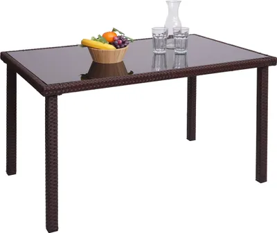 A rectangular poly-rattan table with a glass top, set against a white background. A bowl of fruit, two glasses, and a carafe are placed on the table.
