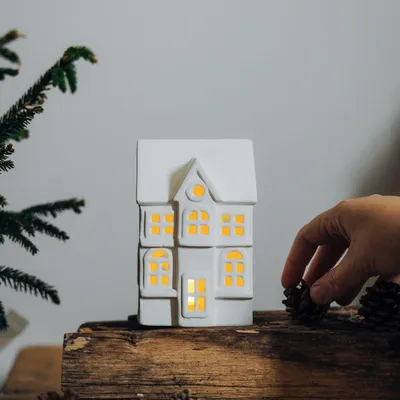A white ceramic house decoration with illuminated windows sits on a wooden surface next to a Christmas tree branch. A hand is placing a pinecone near the house.