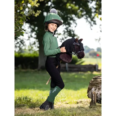 A young girl in equestrian attire holds a hobby horse in a grassy outdoor setting.