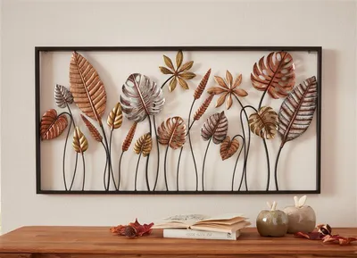 A metal wall art piece featuring various leaf designs is displayed on a wall above a wooden table with books and decorative apples.