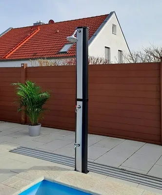 An outdoor shower is set up next to a swimming pool, with a wooden fence and a house in the background.
