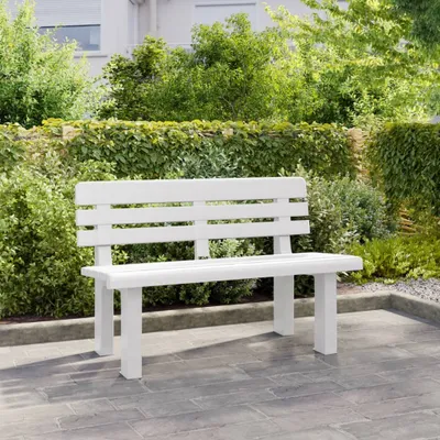 A white garden bench is placed on a paved patio in front of lush green bushes and a building.