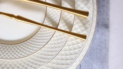 A close-up view of stacked white plates with gold trim and a diamond pattern, with gold cutlery resting on top.