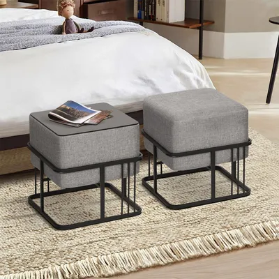 Two gray upholstered ottomans with black metal frames are placed on a textured rug next to a bed.