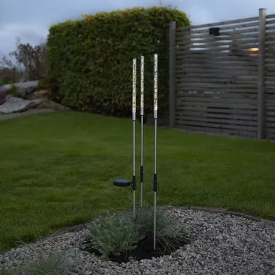 Three LED solar stakes are planted in a garden bed with gravel and low-lying plants. A wooden fence and a hedge are in the background.