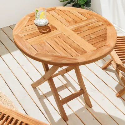 A round, wooden garden table with a glass of drink on it, placed on a wooden deck.