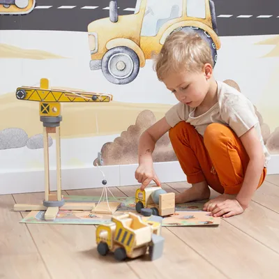 A young boy is playing with a wooden construction toy set on the floor, which includes a crane and trucks. The background features a mural of construction vehicles.