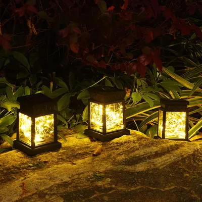 Three solar-powered lanterns with LED string lights are placed on a rocky surface in a garden at night.
