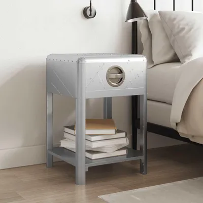 A silver bedside table with a drawer and a shelf is placed next to a bed. Books are stacked on the shelf.