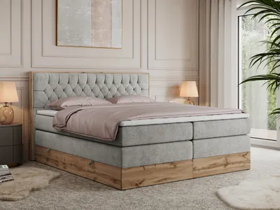 A bedroom scene featuring a large boxspring bed with a tufted headboard and a wooden base, set against a wall with decorative molding and a large window with sheer curtains.