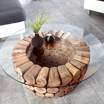A round coffee table with a glass top and a base made of stacked wood logs, placed on a tiled floor next to a sofa.