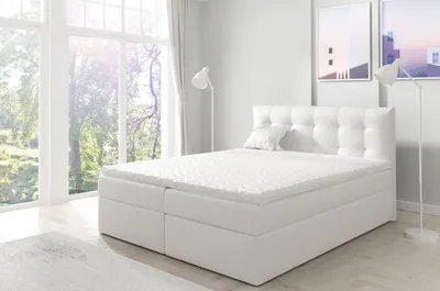 A white upholstered boxspring bed with a mattress topper is shown in a bright, modern bedroom with large windows.