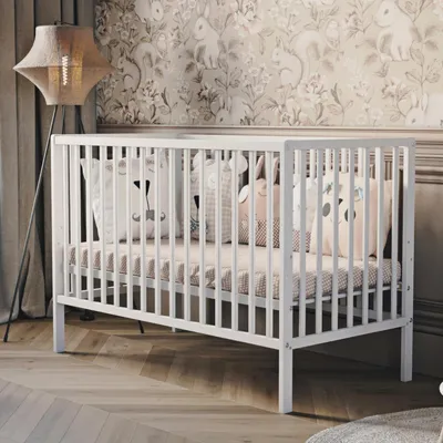 A white baby crib with decorative pillows inside, set in a nursery with patterned wallpaper and a floor lamp.