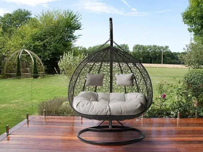 A hanging egg chair for two people is placed on a wooden deck in a garden setting.