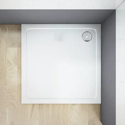 A top-down view of a white square shower tray with a drain cover in the corner, set against a wooden floor and gray walls.