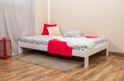 A white wooden bed with patterned bedding, red and cream pillows, a red blanket, and a white fluffy throw is shown in a room with wooden floors and a sheer curtain.