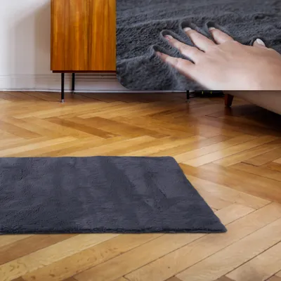 A dark gray shaggy rug is placed on a wooden floor in front of a mid-century modern wooden cabinet. A hand is shown pressing into the rug, highlighting its softness.
