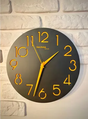 A black analog wall clock with orange numbers and hands, mounted on a white brick wall.