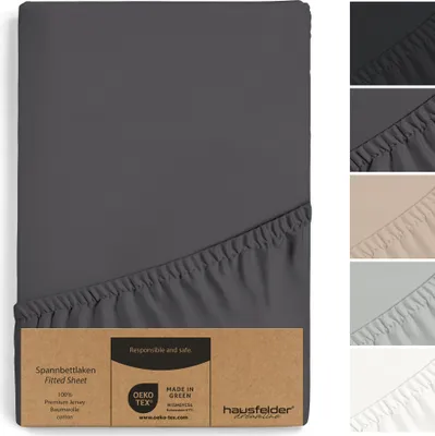 A fitted sheet is shown folded with a cardboard tag attached. Several swatches of different colored fitted sheets are displayed on the right side of the image.