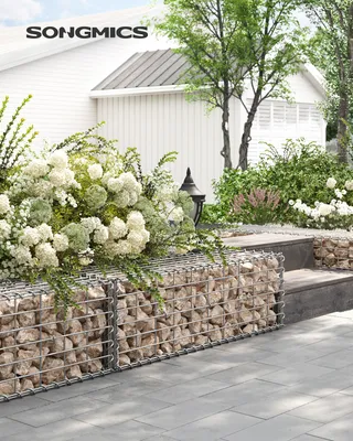 A gabion basket filled with stones and topped with white flowers, placed in a garden setting next to a white house.