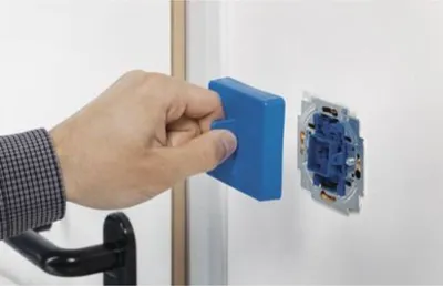 A hand is holding a blue tool and is about to place it onto a wall-mounted electrical socket.