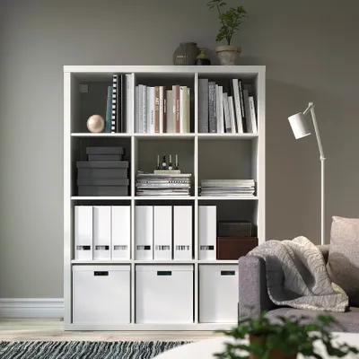 A white shelving unit filled with books and storage boxes, placed in a living room setting next to a sofa and a floor lamp.
