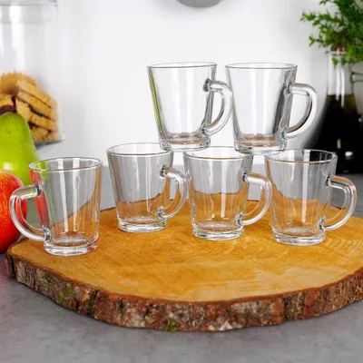 A set of six clear glass espresso cups arranged on a wooden board, with some fruit and cookies in the background.
