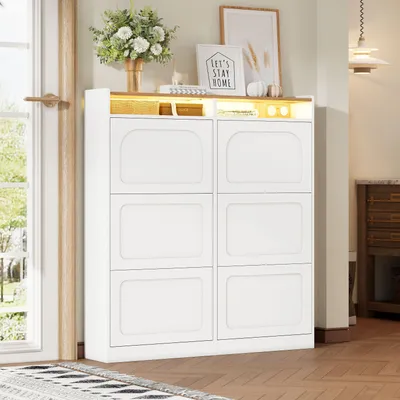 A white shoe cabinet with six flip-down doors and an open shelf with LED lighting on top, decorated with a vase of flowers and framed pictures.