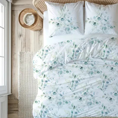 A top-down view of a bed with eucalyptus-patterned bedding, a woven basket with a hat, and a rug on a wooden floor.