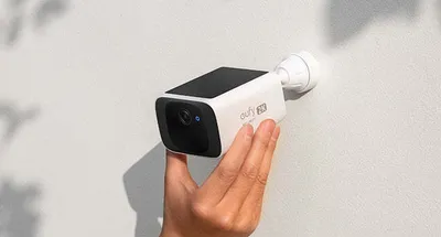 A hand is holding a white and black Eufy security camera against a textured white wall, with shadows of leaves visible on the wall.