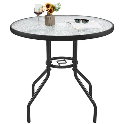 A round glass-top table with a black metal base, set against a white background. A wine glass, a vase with flowers, and sunglasses on a piece of paper are placed on the table.