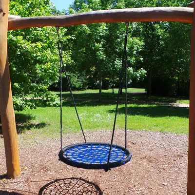 A blue nest swing hanging from a wooden frame in a park-like setting with trees and grass.