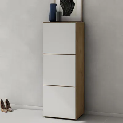 A tall, narrow cabinet with three white doors and a wood-grain side panel, standing against a lightly textured gray wall. A pair of shoes is visible on the floor to the left.