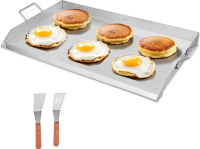 A stainless steel griddle with pancakes and fried eggs on it, along with two spatulas below.