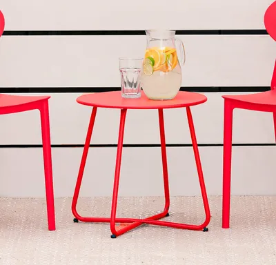 A red metal side table with a pitcher of infused water and a glass on top, placed outdoors next to two red chairs.