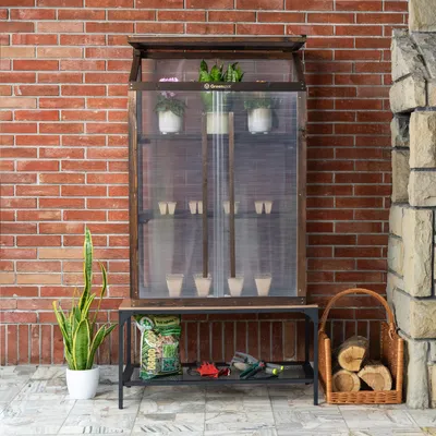 A mini greenhouse on a stand next to a brick wall, with gardening supplies and firewood nearby.