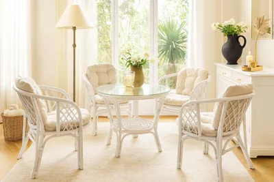 A white rattan dining set with a glass-top table and four chairs is arranged in a bright, sunlit room with large windows.