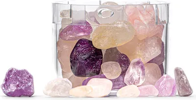 A clear glass container filled with colorful polished stones, with some stones scattered in front of the container on a white surface.