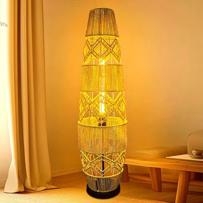 A tall, woven floor lamp with a light bulb inside, standing next to a beige curtain and a wooden bench.
